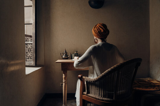 Elegant woman in turban sitting at table in room