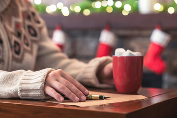 Woman writing letter to santa claus using fountain pen on sheet of paper at christmas fireplace with decoration of light bulbs drinking hot cocoa and marshmallow.