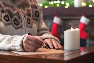 Woman writing wish list using fountain pen on sheet of paper at christmas fireplace with decoration of light bulbs and candle on table.
