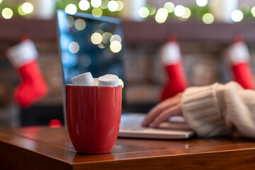 Lonely woman freelancer enjoying with cup of hot cocoa and marshmallow working on laptop near christmas fireplace with decoration of light bulbs.