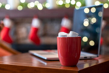 Laptop, cup of hot chocolate with marshmallow on the table at christmas fireplace with decoration of light bulbs. Close up.