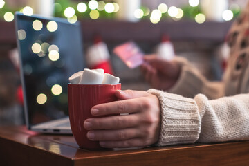 Woman in sweater holding credit card using laptop for making order sitting at table near christmas fireplace with decoration of light bulbs. Online booking, reservation. Travel concept