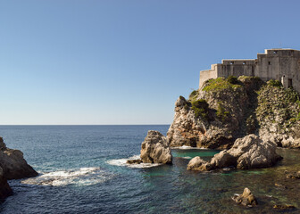 The sea, some rocks and a part of the historic city wall in Dubrovnik, Croatia. The sky is blue.