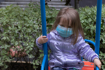 Portrait of a cute blonde little girl wearing a Medical Disposable Face Mask, playing on a playground during quarantine. Stay at home. Isolation. Coronavirus and Air pollution concept.