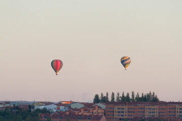 Dos globos aerostáticos flotan sobre la ciudad. Tienen colores vivos. El cielo está despejado. en el paisaje urbano se pueden distinguir edificios residenciales, árboles y generadores eólicos.