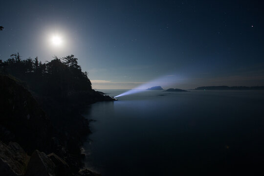 A person shining a bright flashlight into the night sky over the ocean