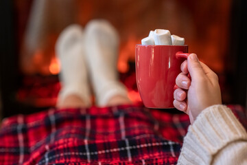 Woman with cup of hot cocoa and marshmallow warming legs in winter white socks near fireplace flame, covered christmas plaid