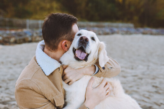 Handsome young man sit on beach and hug and kiss his beautiful golden retriever.