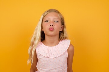Beautiful Caucasian young girl standing against yellow background, keeps lips as going to kiss...