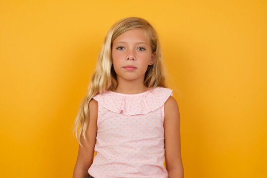 Joyful Beautiful Caucasian young girl standing against yellow background looking to the camera, thinking about something. Both arms down, neutral facial expression.