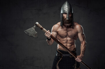 Nord barbarian with naked torso and beard in helmet posing holding his two handed axe in dark studio background.