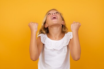 Beautiful Caucasian young girl standing against yellow background looks with excitement up, keeps hands raised, notices something unexpected.