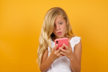 Beautiful Caucasian little girl standing against yellow background looks with bugged eyes, holds modern smart phone, receives unexpected message from friend, reads reminder.