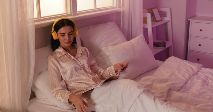 Beautiful young woman listening to music while reading magazine in bed