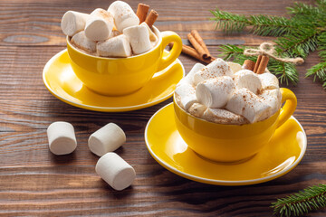 Two yellow ceramic mugs of latte with marshmallows and cinnamon next to spruce branches