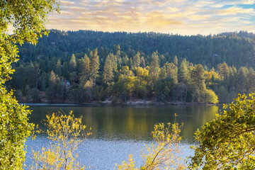 Fototapeta na wymiar a gorgeous shot of the vast still blue lake water with lush green and autumn colored trees reflecting off the lake with majestic mountain ranges at sunset at Lake Gregory in Crestline California