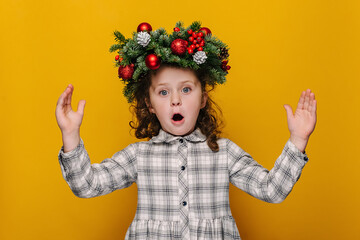 Shocked cute little kid girl showing big size gesture with hands, impressed child demonstrate something wide, wears handmade wreath, isolated on yellow studio background. Merry Christmas concept
