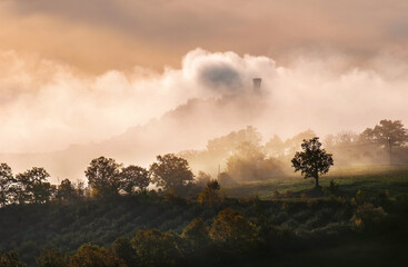 Rocca di Radicofani main tower fortification covered with morning mist. Sunrise light covering the curly clouds running over meadows and fields making light-shadows playing on Tuscany hills landscape.