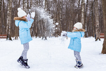 Fototapeta na wymiar Little girls, sisters are walking, having fun in snowy winter park. Stylish clothes, blue jackets with fur,warm pants with snowflakes,cat-shaped mittens.Family picnic in cold weather.Outdoor activity