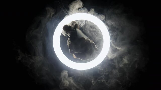 Hand with vape and smoke in slow motion passes through the circle of light on black background. Steam spray from from an electronic cigarette