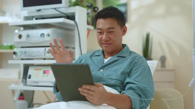 Asian handsome man pating lying in bed at hospital private room videochatting on web screen tablet digital computer with family talking sharing interior communicating. Healthcare. Technology.