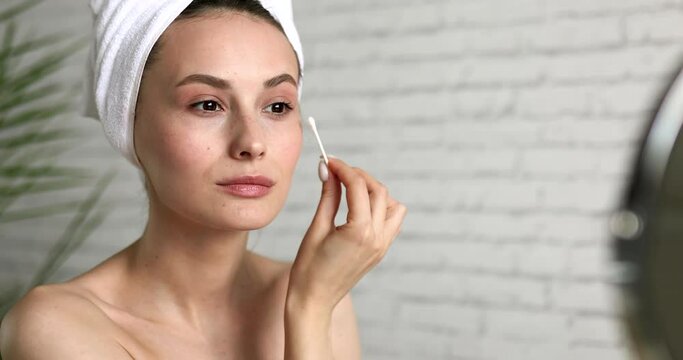 Charming young lady with bath towel on head cleaning skin under eyes with cotton swab. Beautiful woman enjoying morning rituals at home.