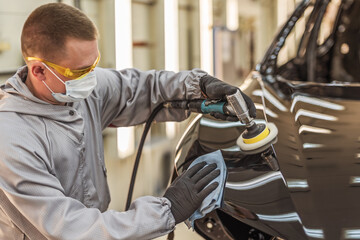 An employee of the car body paint shop with a medical mask on his face polishes the painted surface