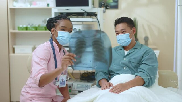African female physician expert with asian patient discussing x-ray image on lungs for diagnosticws in healthcare facilities center hospital indoors.