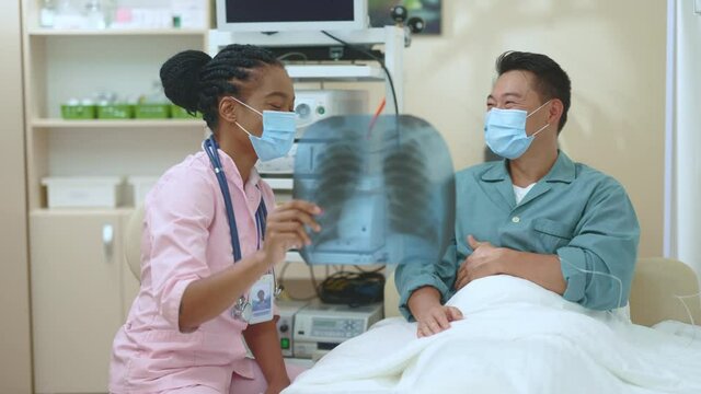 Afro-american female doctor and asian cheerful patient discussing x-ray photograph of lungs claiming poositive diagnosis enjoying talking consulting in hospital room.