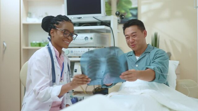 Afro-american female physician discussing lungs fluorography image with adult chinese patient exploring medical checkup for further treatment. Hospital. Clinic.