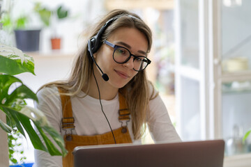 Gardener millennial woman wear headphones using laptop, communicating with clients, watching webinar or video stream conference, remote work from home during lockdown. Distance job, e-business. 