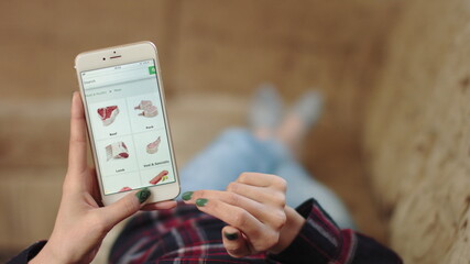 Woman at Home Lying on Couch in Living Room Using Smartphone Buys in Internet Shop. Woman Orders Meat Home In An Online Store Using a Smartphone. Female Selects Meat in Grocery Online Store. POV