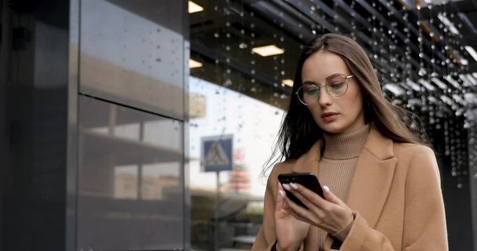 Beautiful stylish female wearing beige coat and eyeglasses walking at city with personal smartphone in hands. Concept of street fashion, technology and lifestyle.