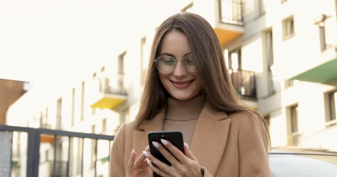 Pleasant young female with natural beauty in beige coat and eyewear using smartphone outdoors. Concept of city lifestyle, people and modern technology.