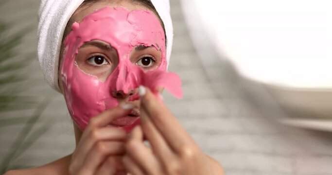 Happy young woman with towel on head taking off pink alginate mask from face while looking at round mirror. Attractive lady using professional cosmetics regularly at home.