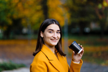 Female hand holding a cup of blank paper on the background of the park. Free space. The girl holds a cup of coffee and drinks coffee. Girl hand hold plastic coffee cup. Woman drinking coffee.