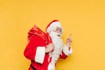 Fototapeta na wymiar Funny Santa isolated on a yellow background with a bag of Christmas presents, looking at the camera and pointing his finger to the side at copy space.