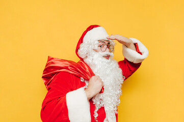 Funny Santa Claus stands on a yellow background with a bag of gifts on his back and poses for the...