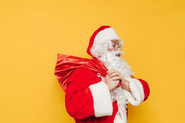 Fototapeta na wymiar Santa Claus with a bag of Christmas presents on his back isolated on a yellow background, looking away. New year concept