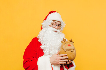 Cute Santa standing on a yellow background with a toy deer in his hands on a pose for the camera at Christmas. New year concept.