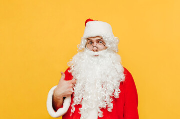 Surprised Santa Claus points a finger at himself and looks at the camera with a shocked face isolated on a yellow background.
