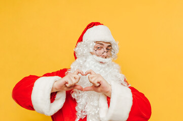 Positive Santa stands on a yellow background and shows his fingers a gesture of love with a smile on his face. Christmas concept.
