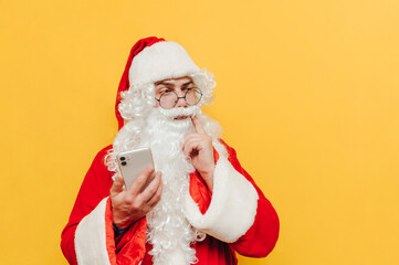 Pensive Santa Claus with a smartphone in his hands stands on a yellow background and looks away at the copy space and thinks. Christmas and New Year