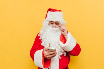 Fototapeta na wymiar Santa Claus enjoys a smartphone on a yellow background with a pensive face at Christmas. Isolated. Christmas concept.