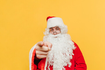 Serious Santa stands on a yellow background, looks at the camera and shows his finger. Santa points a finger at you