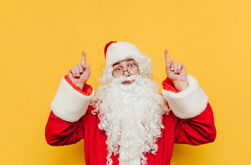 Funny Santa Claus showing thumbs up on copy space with serious face looking at camera, isolated on yellow background. Christmas concept.