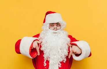 Serious man in santa costume stands on yellow background, looks at camera and shows thumbs down on copy space. Christmas and New Year