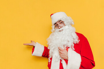 Fototapeta na wymiar Positive Santa Claus stands on a yellow background, holding a copy space and looking at the camera with a smile on his face. Christmas and New Year.
