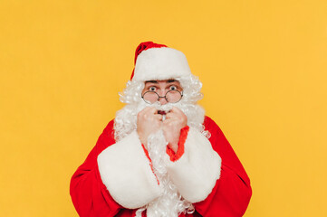 Portrait of shocked santa claus on yellow background, looking at camera with arms raised to mouth, isolated.