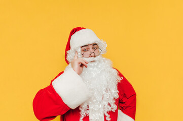 A smart Santa Claus looks at the camera and shows a "think about it" gesture touching his finger to his temple, isolated on a yellow background.
